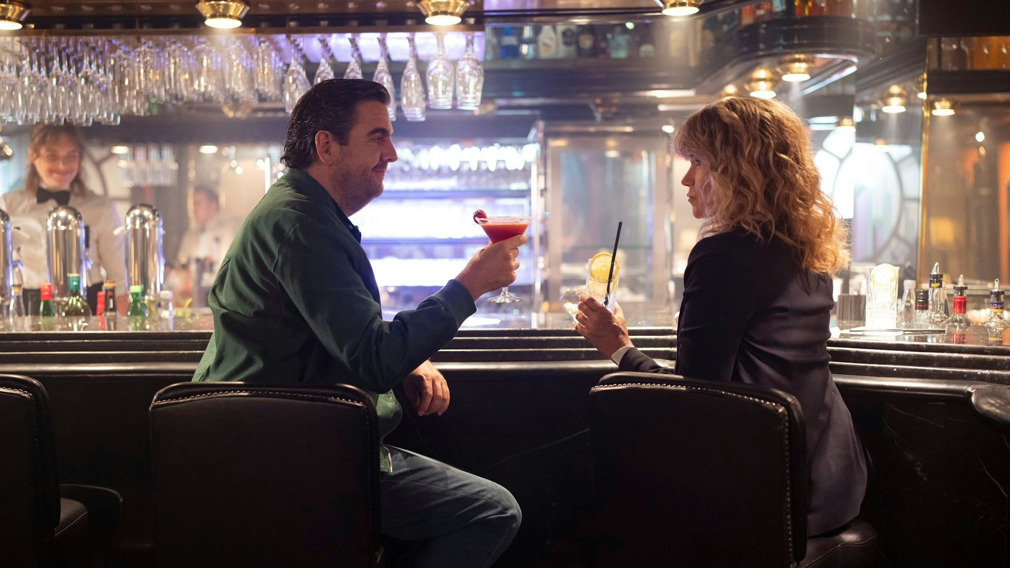 Bastian Pastewka und Anke Engelke prosten sich an einer Hotelbar in ihren als Ralf und Maria in der Serie „Perfekt Verpasst“ zu.