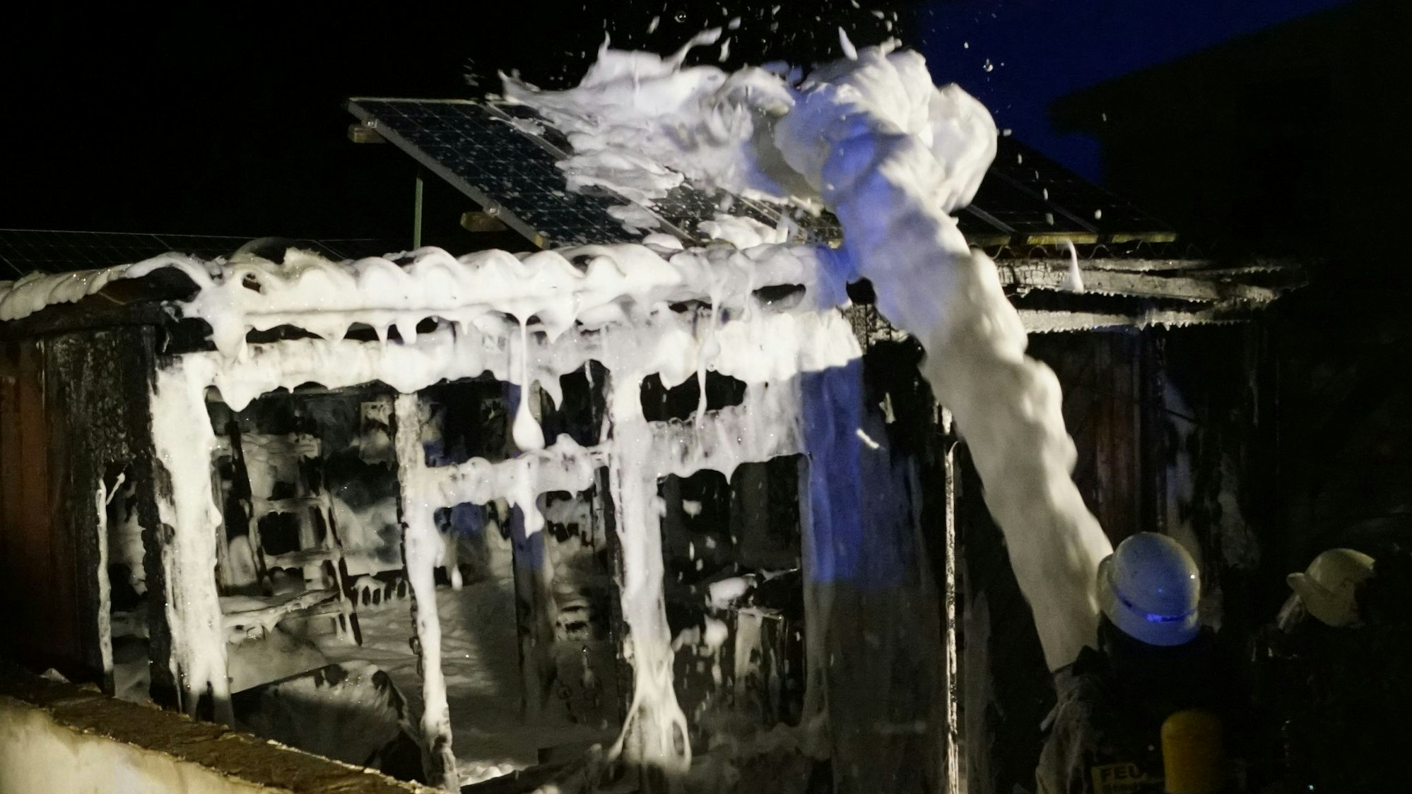 Feuerwehrleute spritzen Schaum auf die Reste der ausgebrannten Gartenlaube.