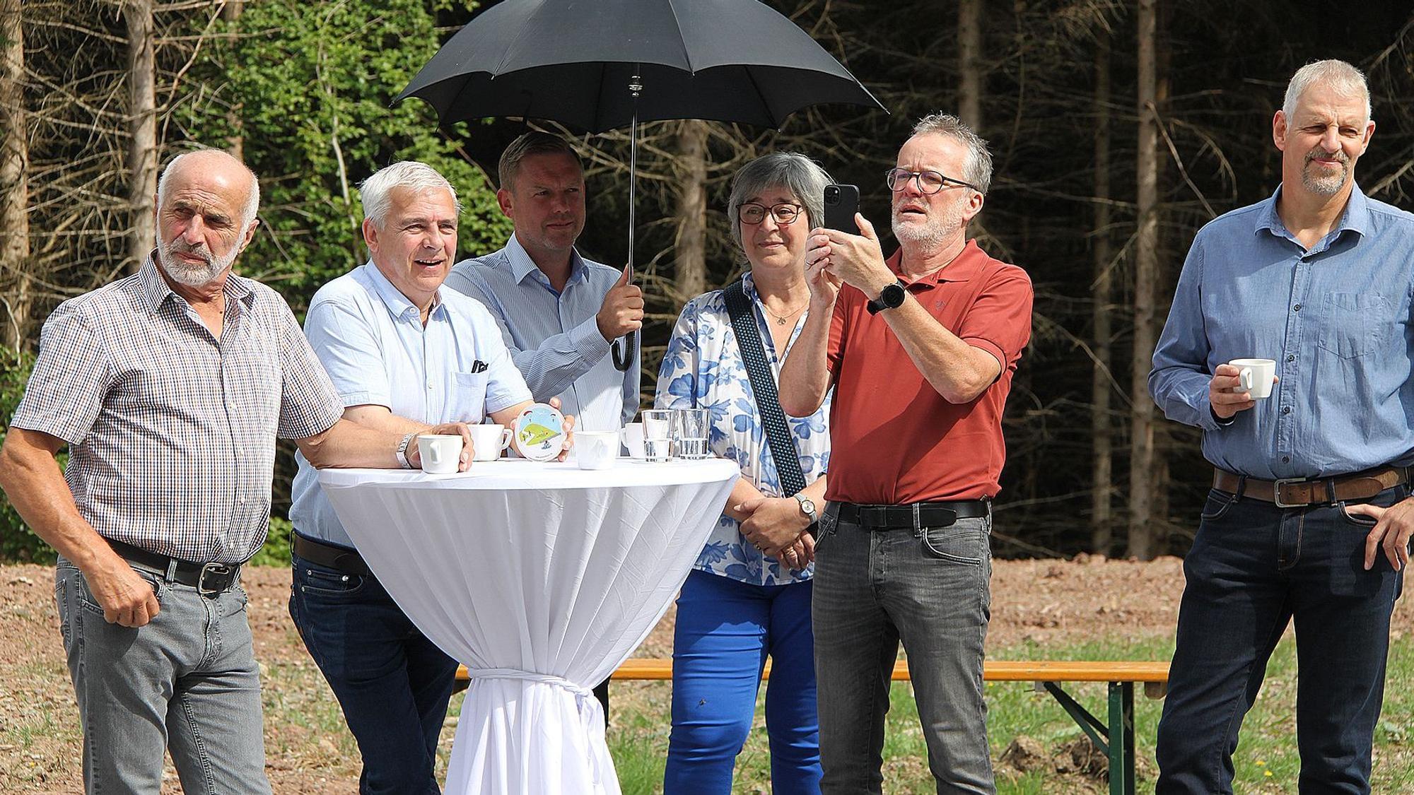 Fünf Männer und eine Frau stehen an einem mit einer weißen Husse bedeckten Stehtisch. Ein Mann hält einen Regenschirm als Sonnenschutz in der Hand.