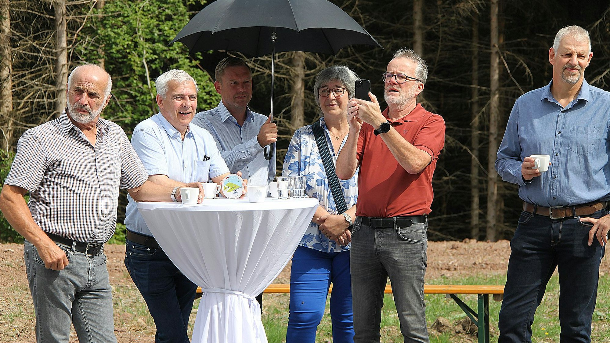 Fünf Männer und eine Frau stehen an einem mit einer weißen Husse bedeckten Stehtisch. Ein Mann hält einen Regenschirm als Sonnenschutz in der Hand.