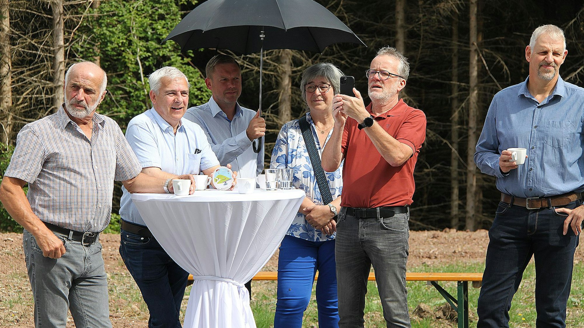Fünf Männer und eine Frau stehen an einem mit einer weißen Husse bedeckten Stehtisch. Ein Mann hält einen Regenschirm als Sonnenschutz in der Hand.