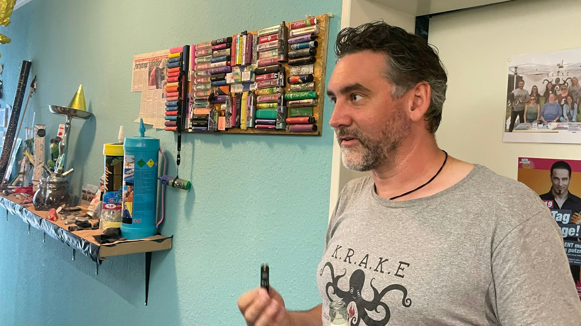 Ein Mann steht an einer Wand, an der allerlei Kleinkram hängt.