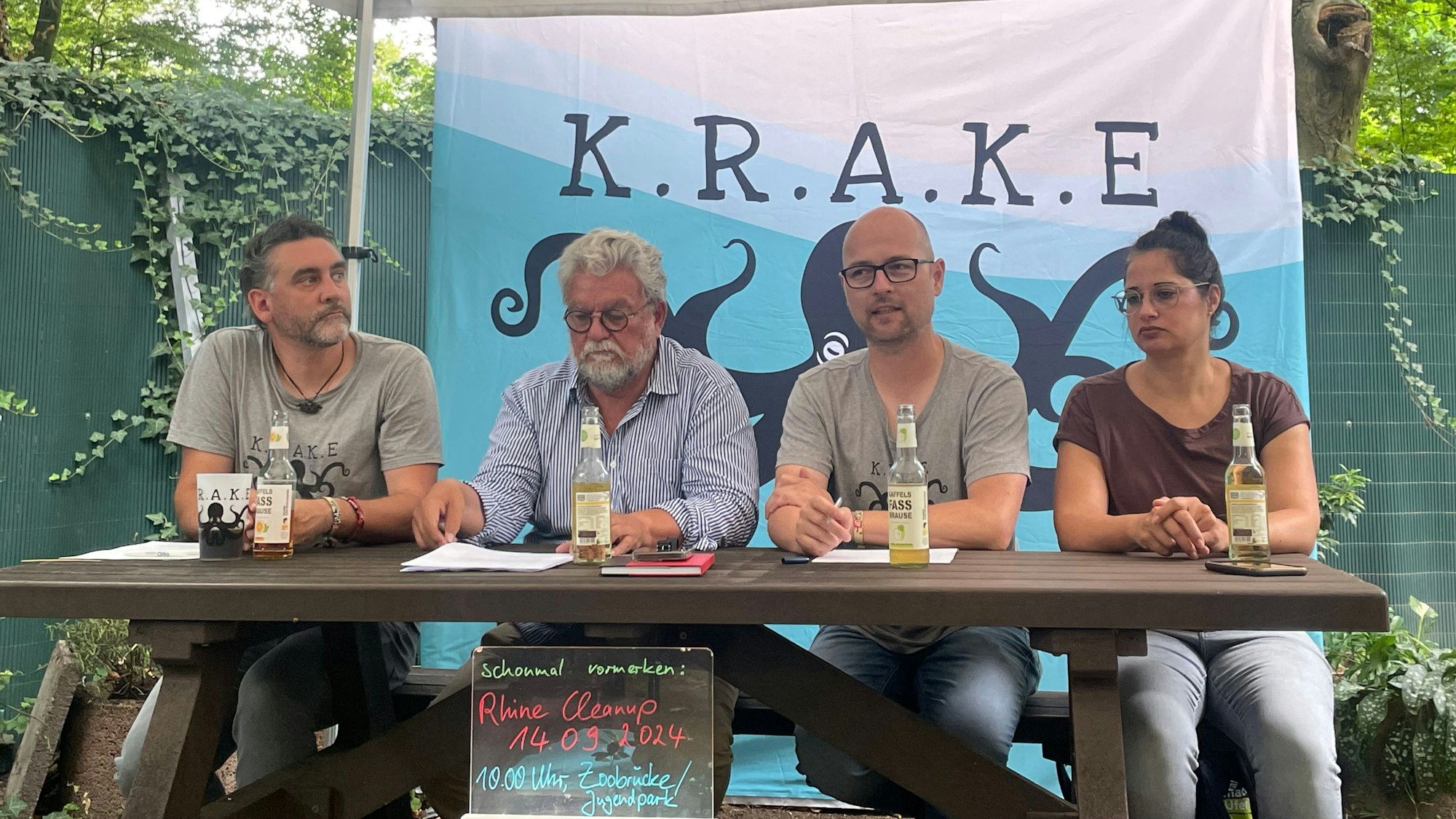 Vier Personen sitzen nebeneinander an einem Tisch, im Hintergrund ist ein Plakat, auf dem K.R.A.K.E steht.