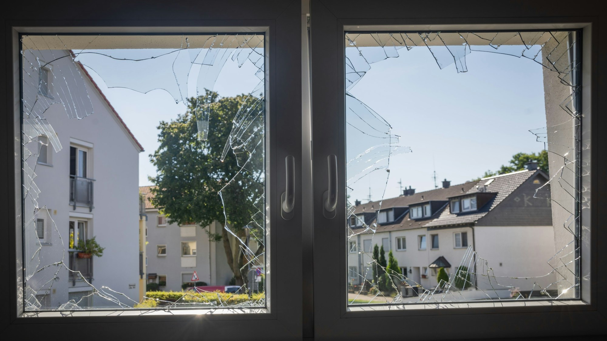 Zu sehen ist ein Doppelfenster mit zerborstenen Scheiben. Dahinter zeigt sich die weitere Wohnbebauung.