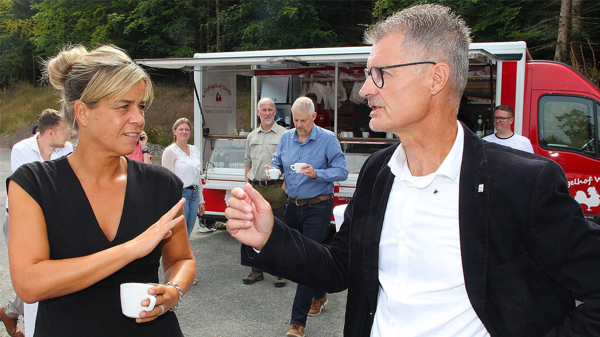 Die stellvertretende NRW-Ministerpräsidentin Mona Neubaur hält eine weiße Kaffeetasse in der Hand und unterhält sich mit Dahlems Bürgermeister Jan Lembach.