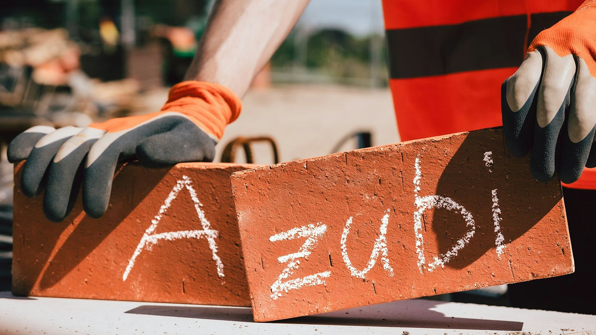 Das Bild zeigt zwei Hände, die zwei Ziegelsteine halten, auf denen das Wort Azubi steht.