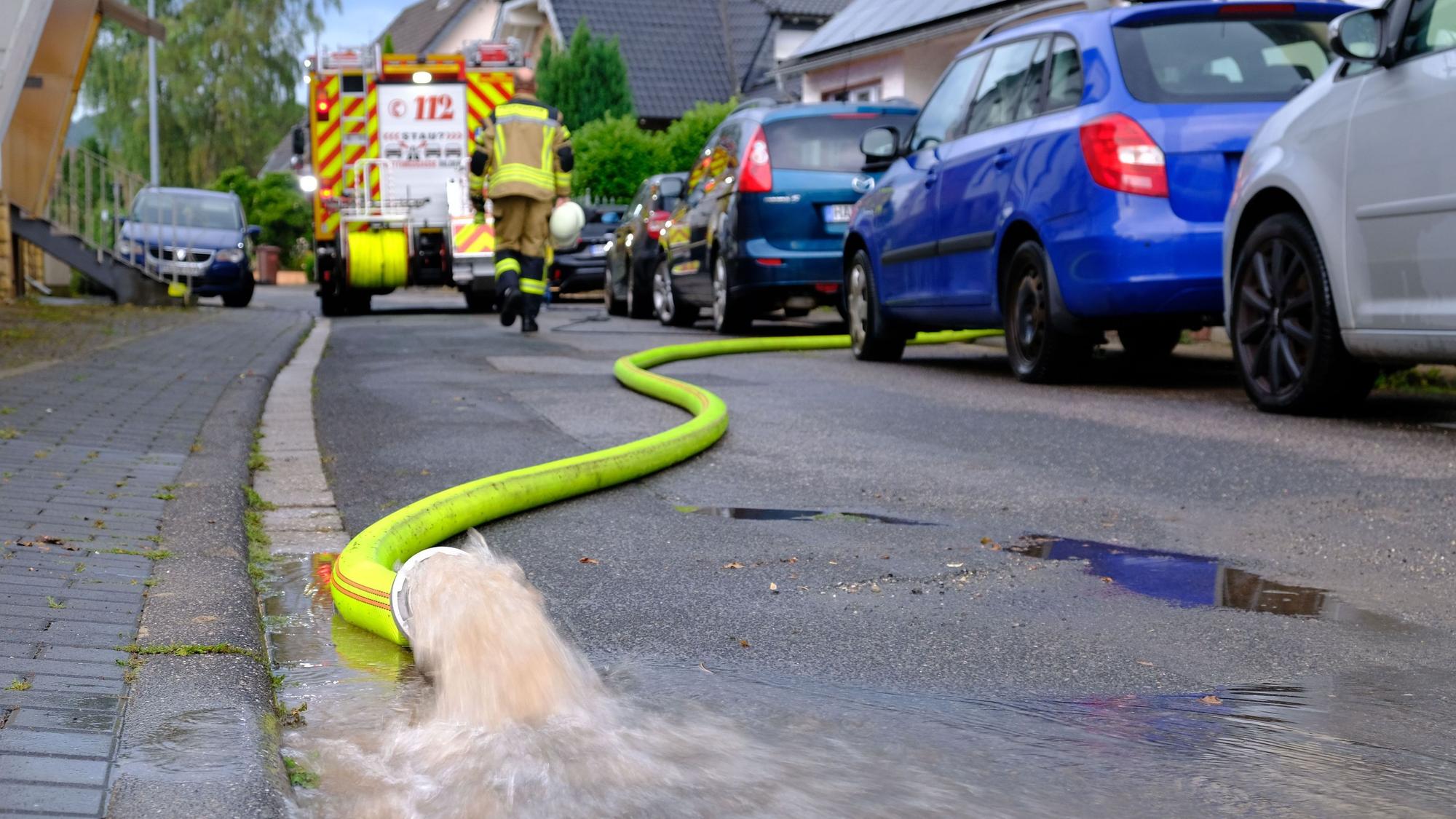 Wasser sprudelt aus einem gelben Feuerwehrschlauch: Helfer der Freiwilligen Feuerwehr pumpen nach einem Starkregenereignis am 13.8.24 Keller in der Rehgasse in Kommern aus.