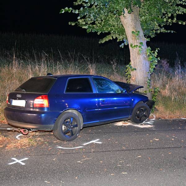 Ein blaues Auto steht kaputt nach einem Unfall an einem Baum. Auf dem Boden Kreidemarkierungen der Polizei.