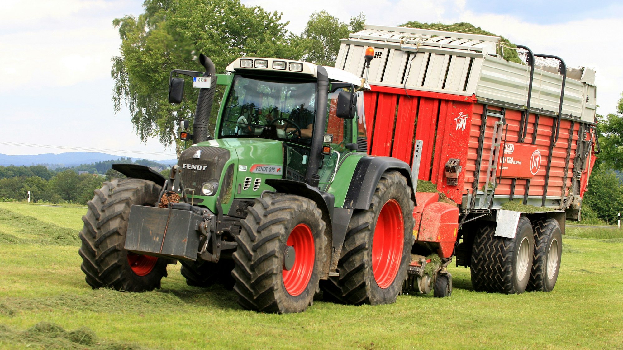 Ein Traktor mit Ladewagen fährt über eine Grünfläche und sammelt Silage ein.