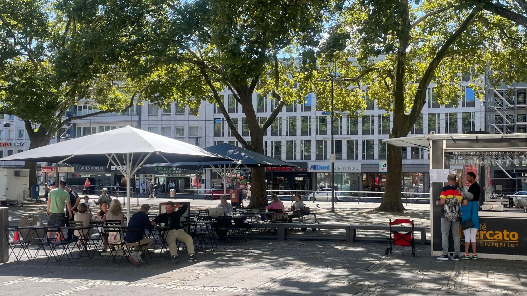 Menschen sitzen auf dem Neumarkt im neu eröffneten Café im Schatten von Platanenbäumen