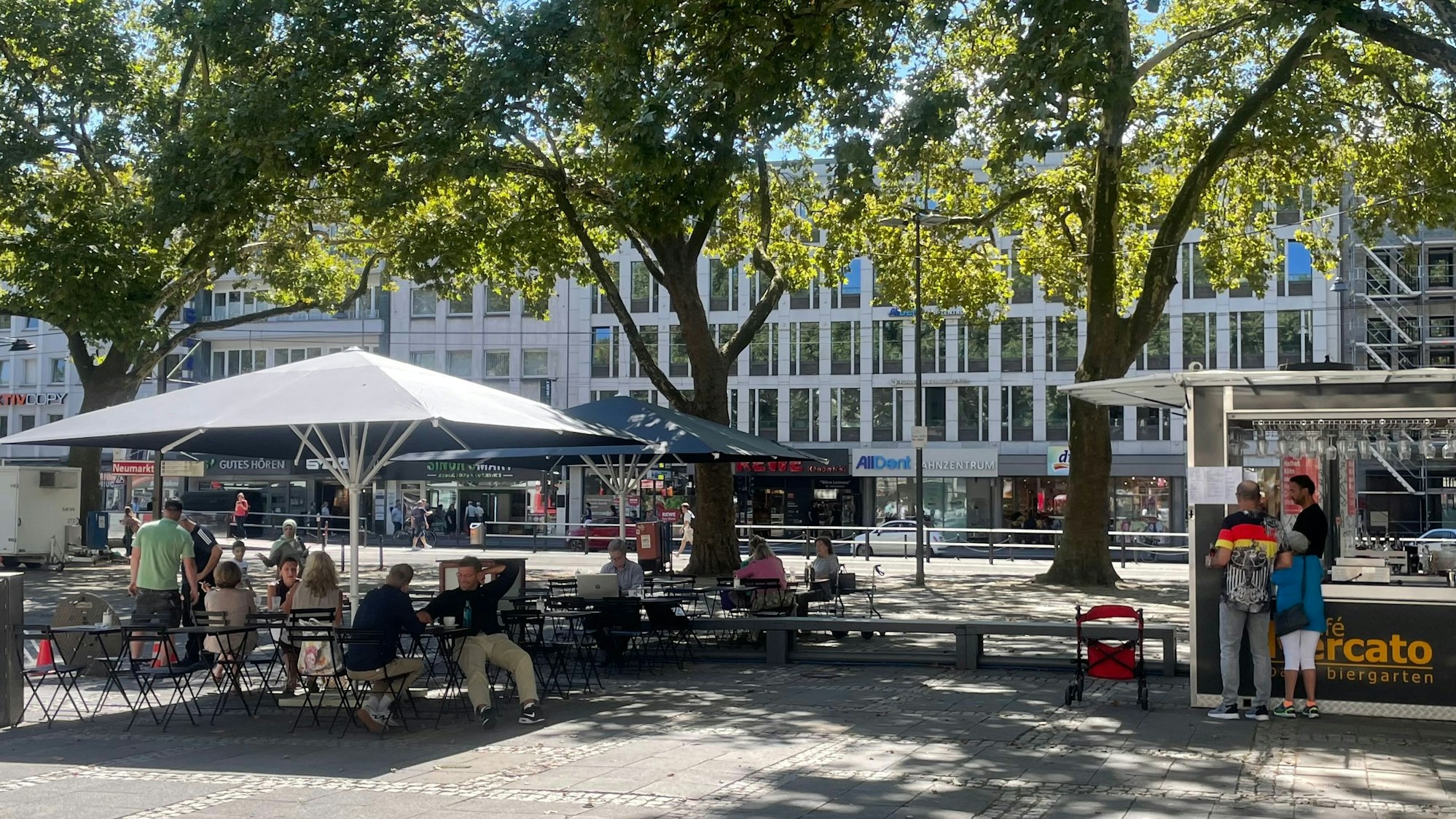 Menschen sitzen auf dem Neumarkt im neu eröffneten Café im Schatten von Platanenbäumen
