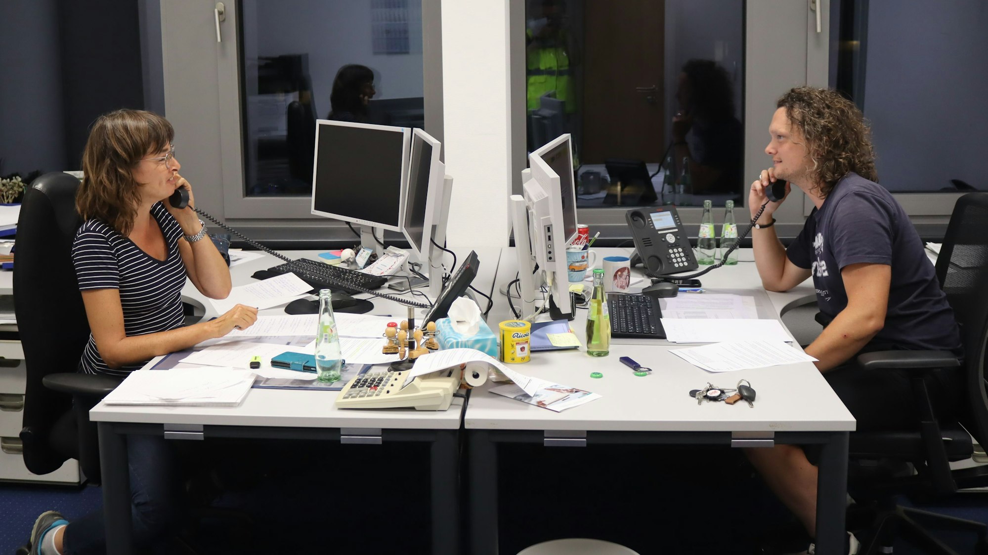 Petra Greif und Björn Schäfer sitzen sich an zwei Schreibtischen gegenüber. Beide telefonieren.