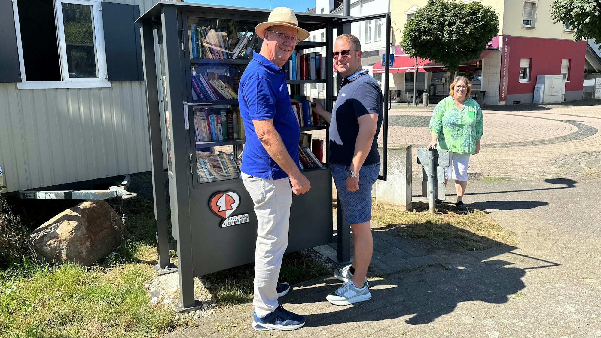 Ortsvorsteher Wilfried Schwab (l.) und Jens Nolden (r.) vom Bürgerverein Hangelar am Bücherschrank am Platz Op d'r Dränk. Rechts außen Natascha Brecht-Kalus vom Werbekreis Hangelar.