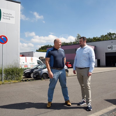 Das Foto zeigt zwei Männer und im Hintergrund zwei Hallen der Firma SN.