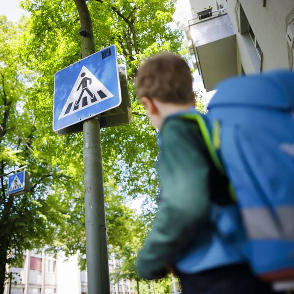 Symbolfoto zum Thema Schulkind im Straßenverkehr. Ein sechsjähriger Junge steht mit seinem Schulranzen auf dem Rücken vor dem Schild eines Fußgängerüberweges.