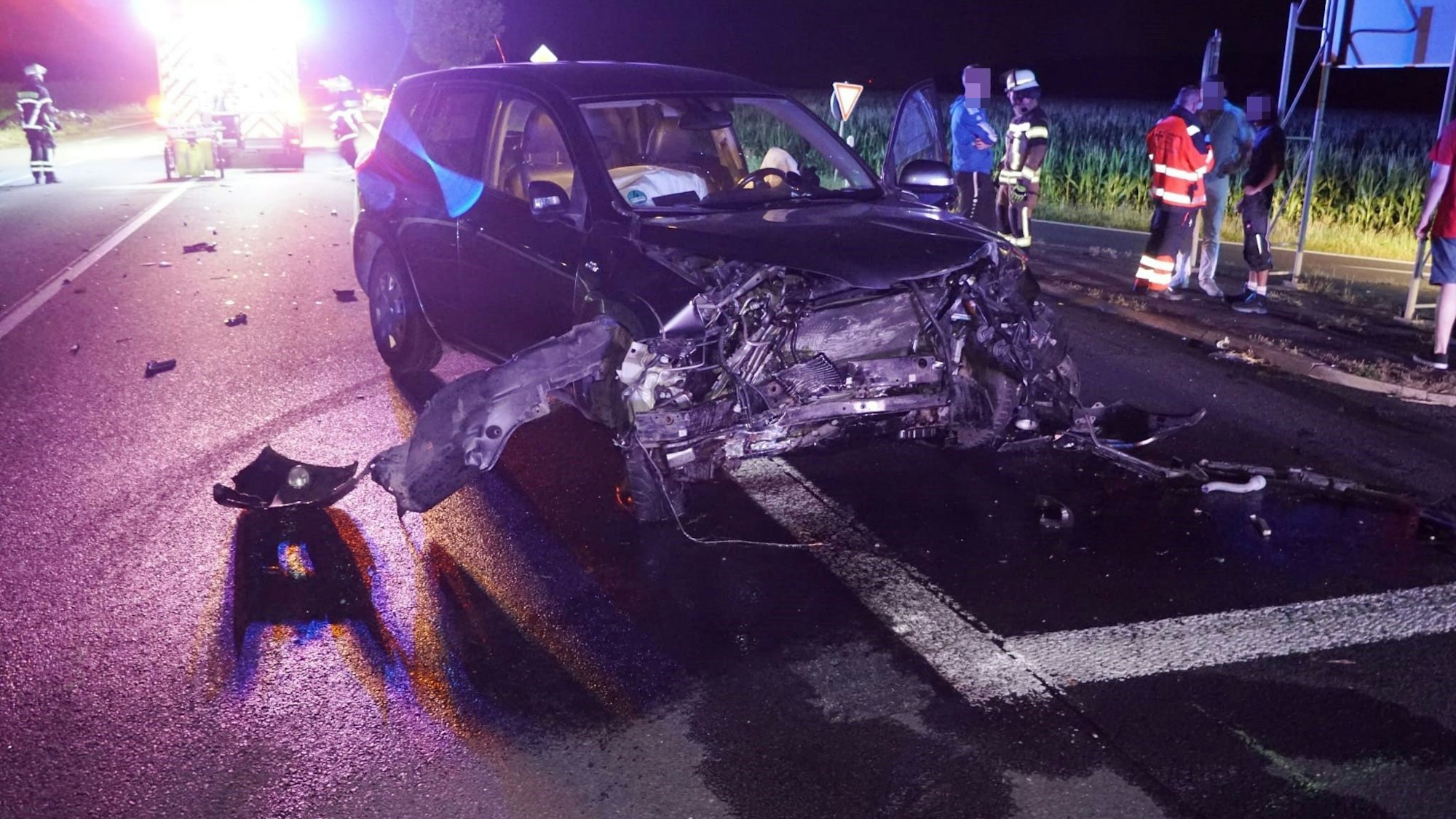 Ein Auto mit demolierter Front steht auf der Straße, im Hintergrund sind Feuerwehrleute.