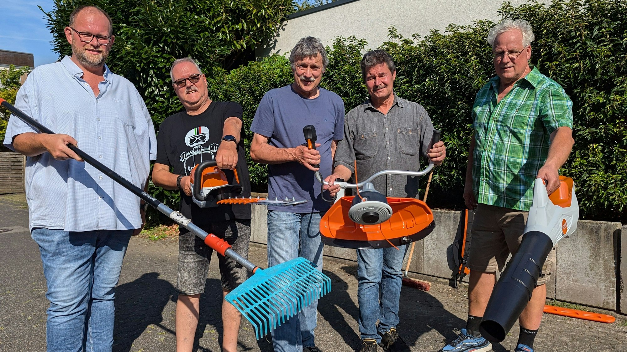 Das Foto zeigt eine Gruppe von Männern, die Gartengeräte wie Harke, Heckenschere, Freischneider und Laubbläser präsentieren.