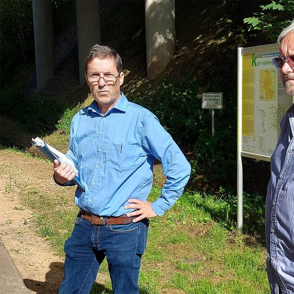 Zwei Männer in blauen Hemden stehen an einem bewaldeten Weg.