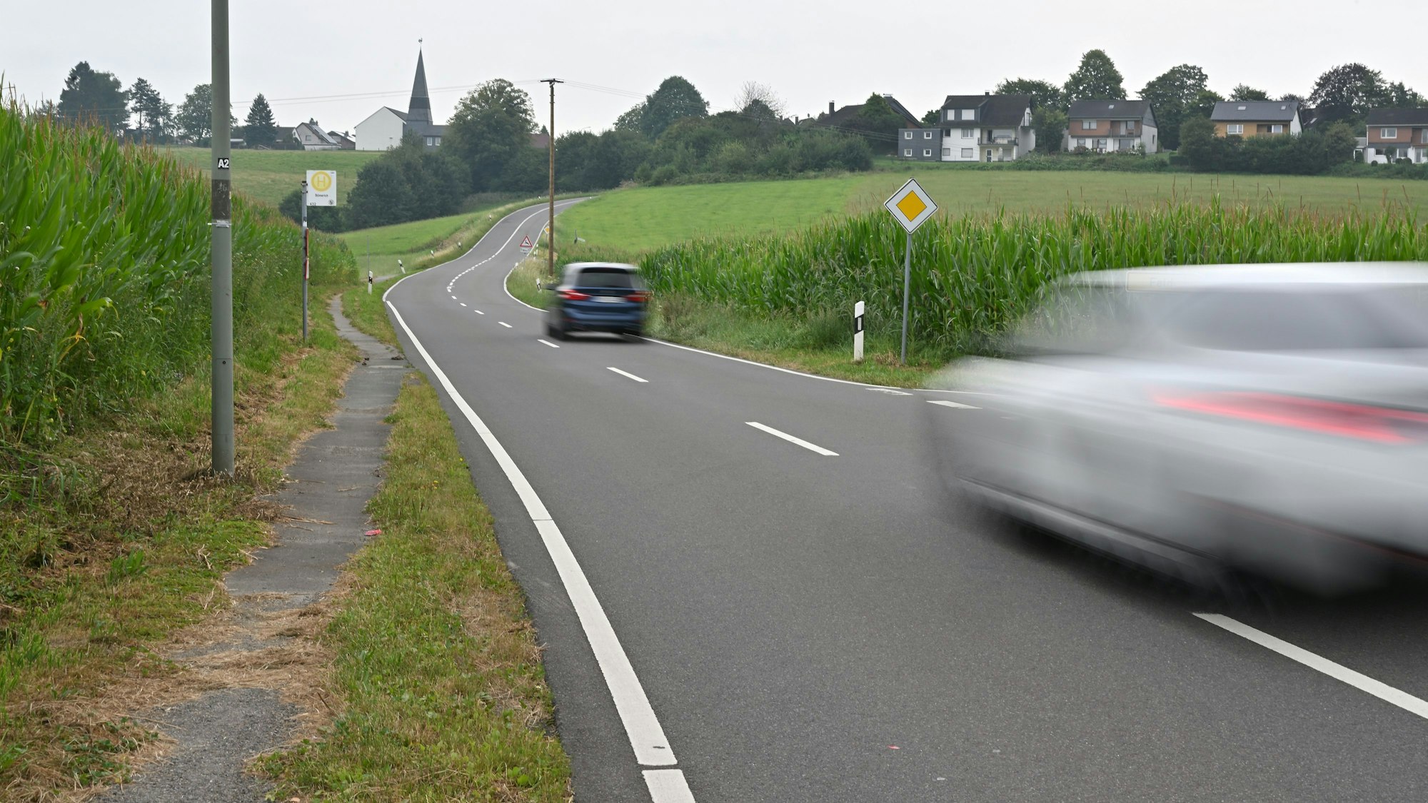 Eine Landstraße. Auf ihr zwei Autos, am Rand nur ein schmaler Trampelpfad für Fußgänger.