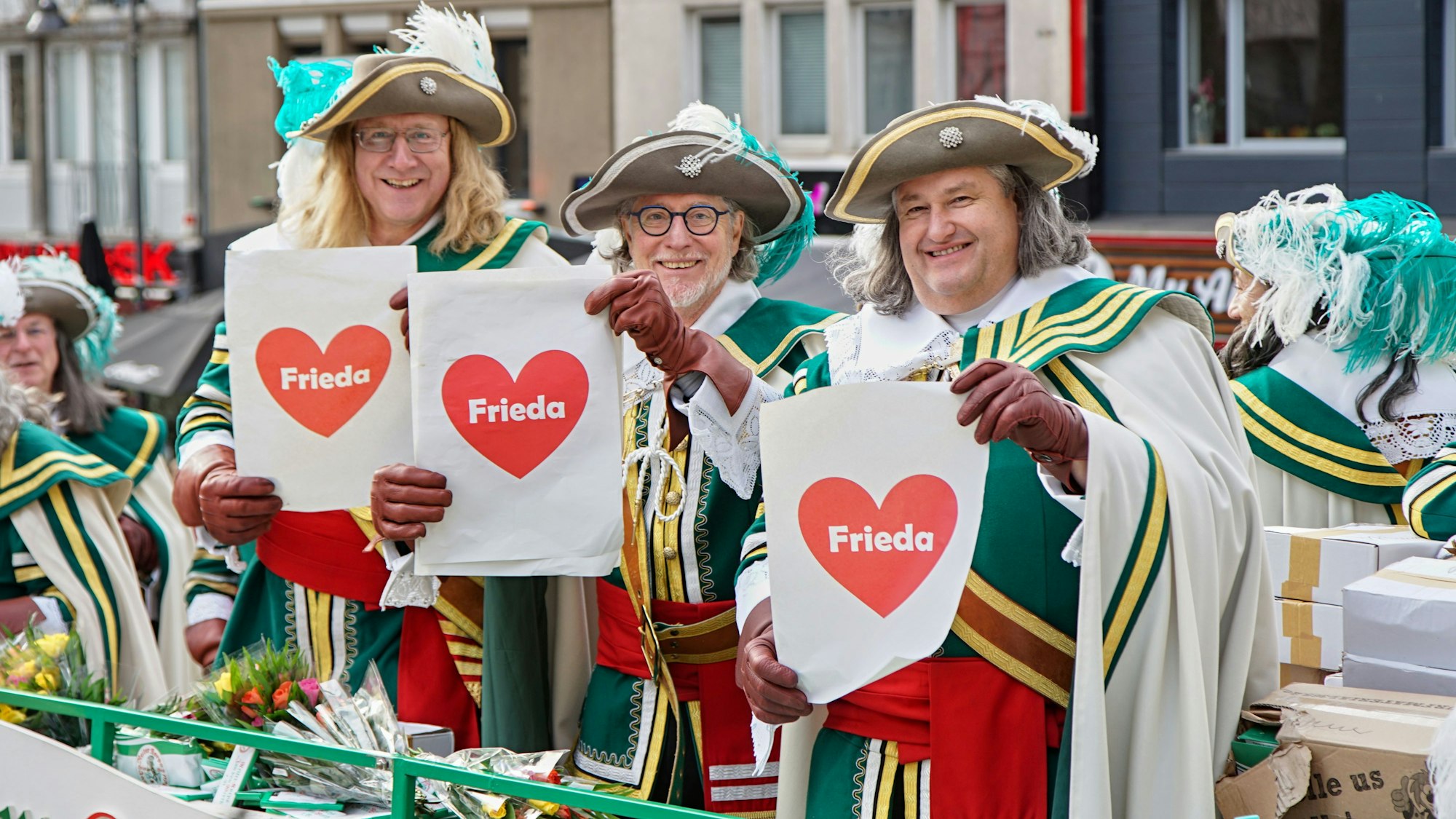 Drei Männer mit Federhut und Blättern, auf denen in einem roten Herz Frieda steht.