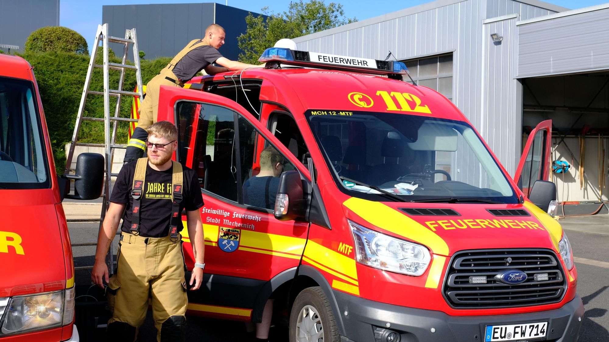 Zwei Feuerwehrleute stehen an einem Mannschaftstransportwagen, auf der Beifahrertür ist das Wappen der Stadt Mechernich zu sehen. Sie bereiten sich darauf vor, die Bevölkerung mit Lautsprecherdurchsagen zu informieren.