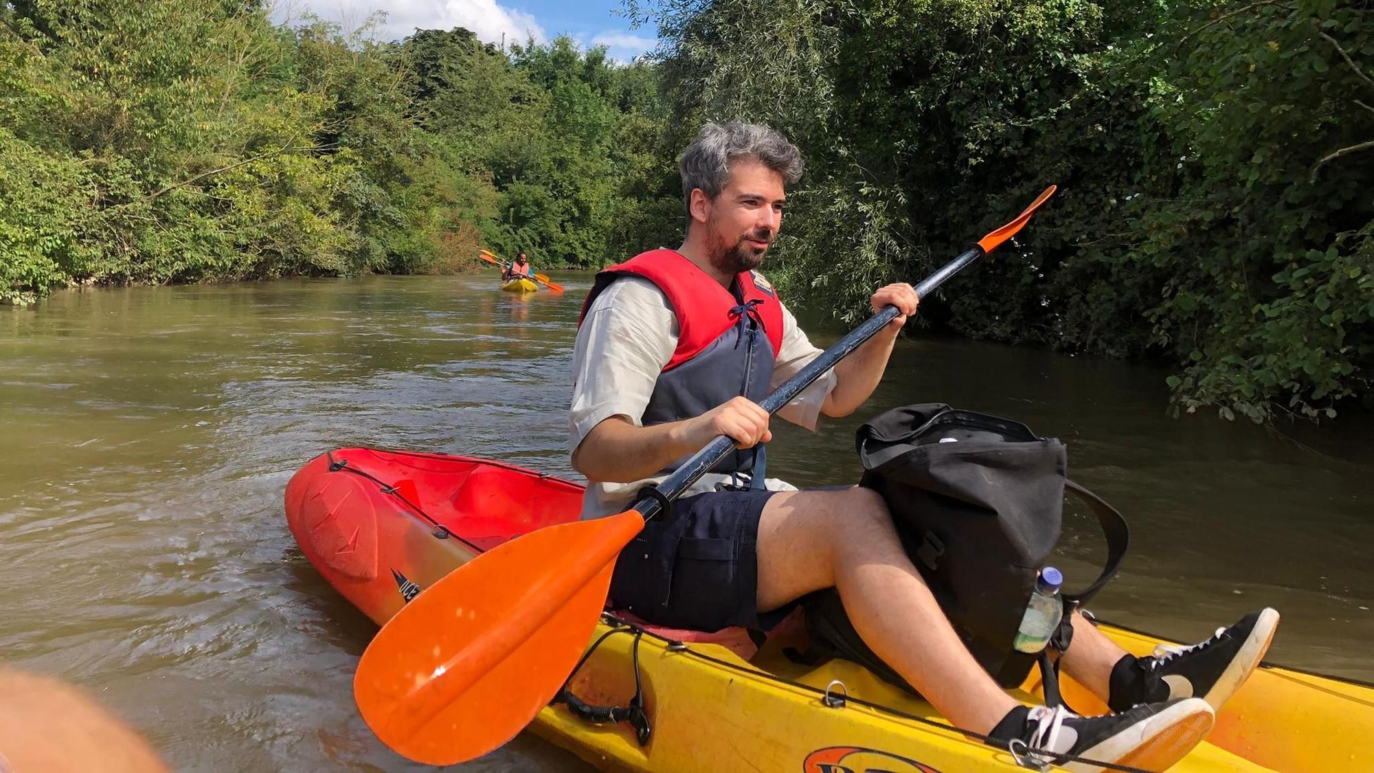 Auf dem Bild ist ein wasserscheuer Journalist zu sehen, der nach bestem Gewissen auf der Erft paddelt. Das Kajak ist vorne gelb und hinten rot. Im Hintergrund Büsche und Bäume am Ufer des Wassers.