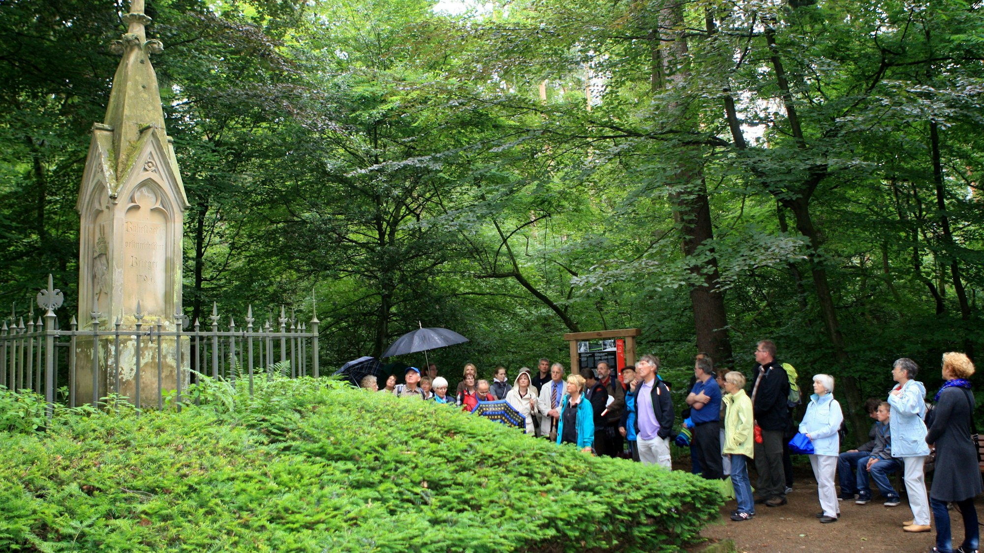 Eine Wandergruppe steht am Denkmal für im Bensberger Schloss verstorbene österreichische und preußische Soldaten mitten im Wald der Hardt.