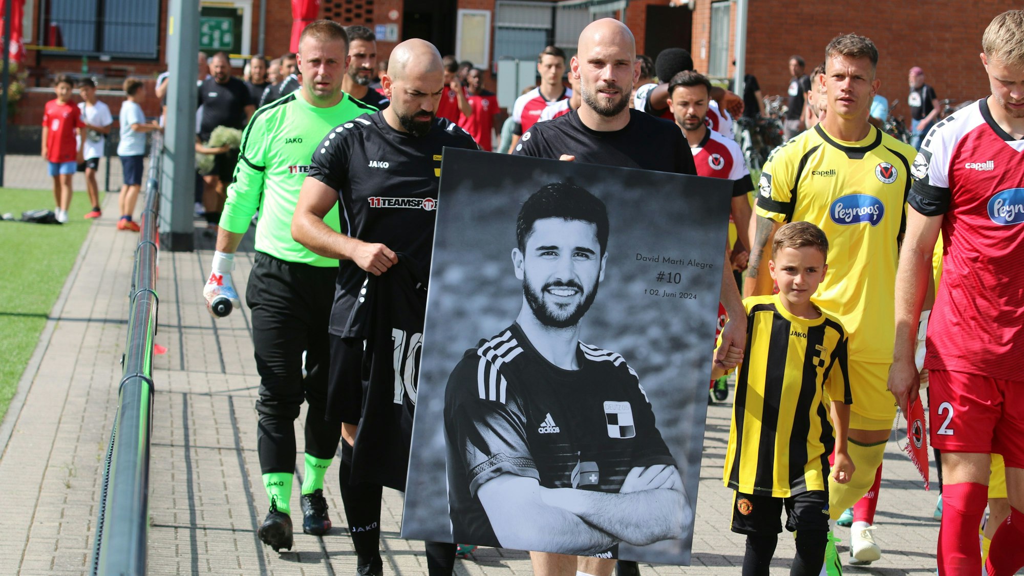 Die Spieler gehen von der Kabine auf den Platz. Einer trägt ein schwarz-weißes Foto des verunglückten Spielers.