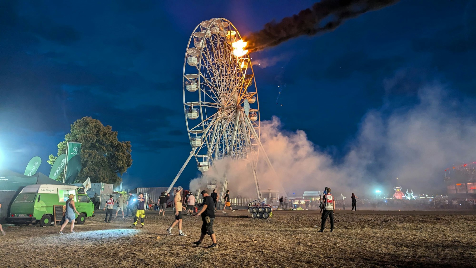 Besucher des Highfield-Festivals und Helfer blicken auf ein Riesenrad, an dem Flammen zu sehen sind. Auf dem Highfield-Festival am Störmthaler See ist ein Feuer an einem Riesenrad ausgebrochen.