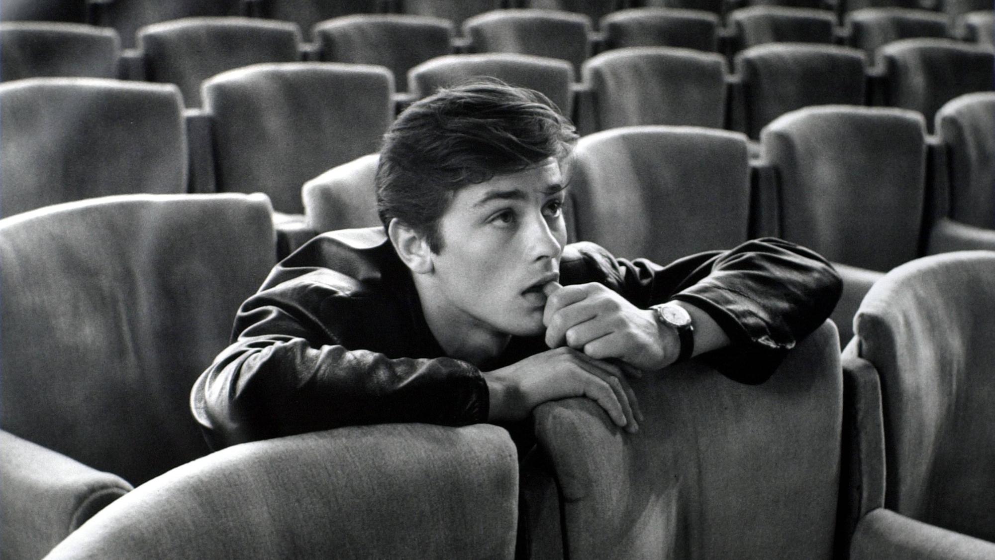 Alain Delon sitzt auf einem Kinosessel und schaut auf die Leinwand.