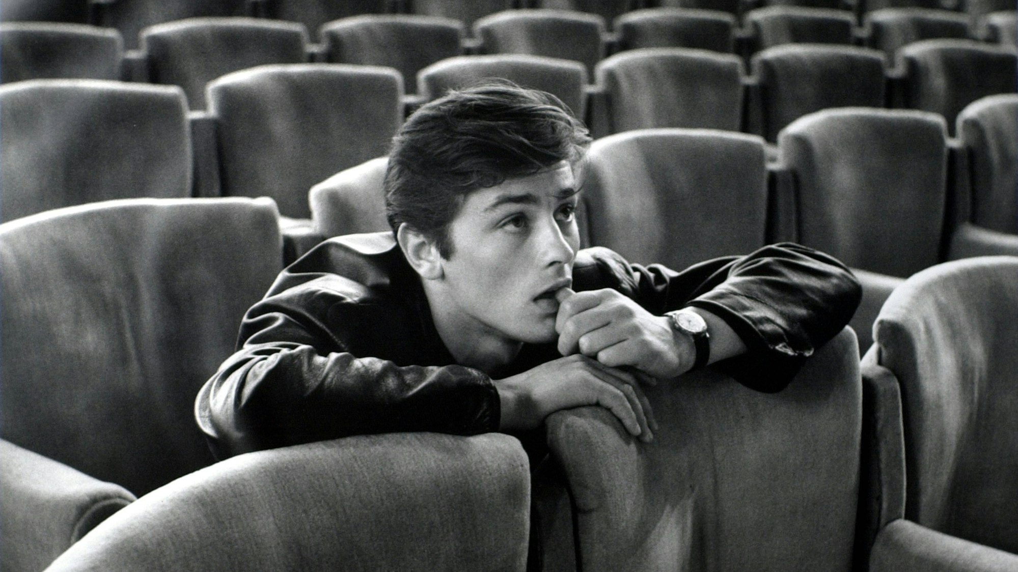Alain Delon sitzt auf einem Kinosessel und schaut auf die Leinwand.