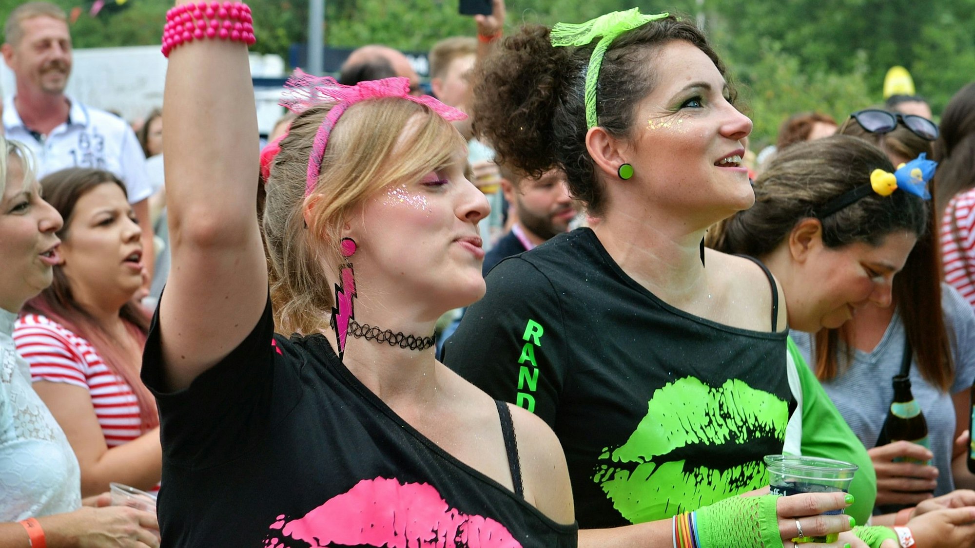 Zwei Besucherinnen des Kölsch-Festivals am Firmenicher Zikkurat. Sie tragen schwarze Shirts und neonfarbene Accessoires.