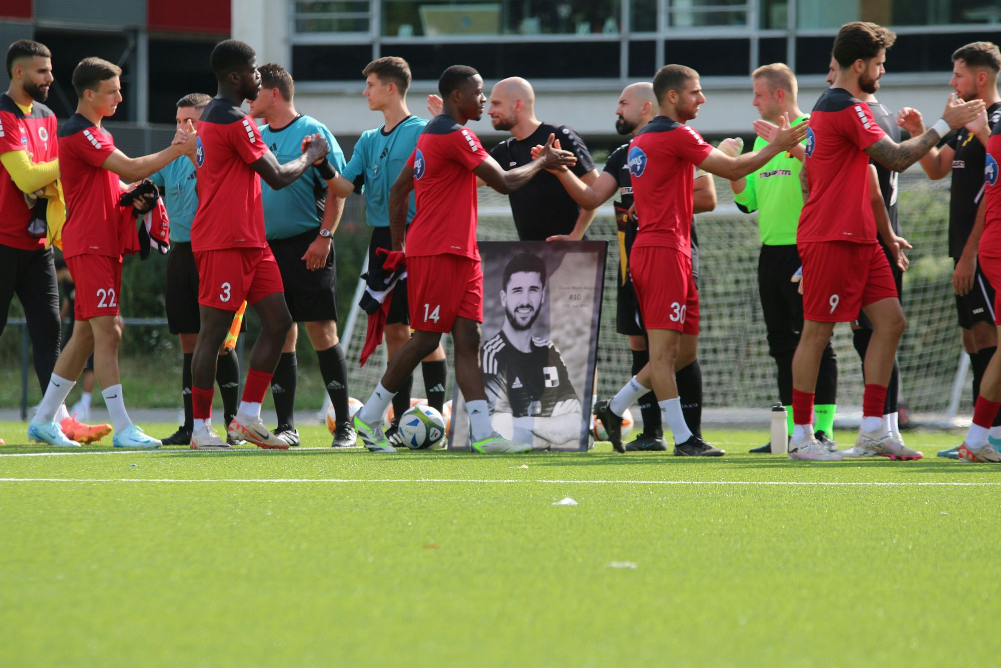 Die Spieler geben sich vor dem Anpfiff die Hand, das Bild von David Marti Alegre steht dabei vor einem der Spieler.