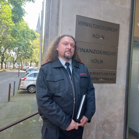 Bild eines Feuerwehrmannes in Uniform vor dem Gebäude des Kölner Verwaltungsgerichtes am Appellhofplatz