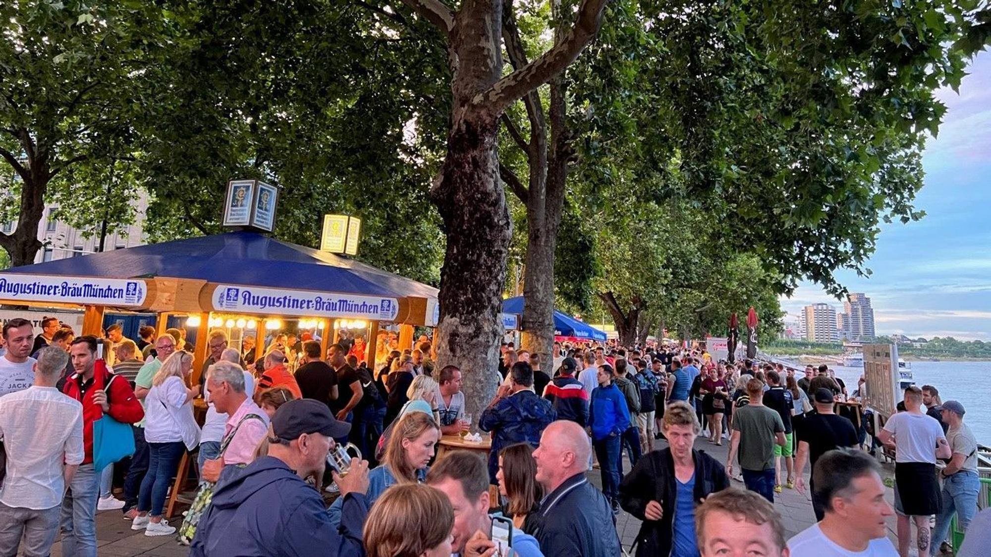 Menschen stehen an der Rheinpromenade an Stehtischen und trinken Bier