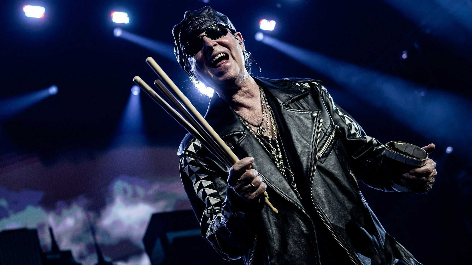 Scorpions-Sänger trägt Sonnenbrille, Leder-Käppi und -jacke auf der Bühne der Wembley-Arena in London. In der rechten Hand hält er mehrere Drum-Sticks.