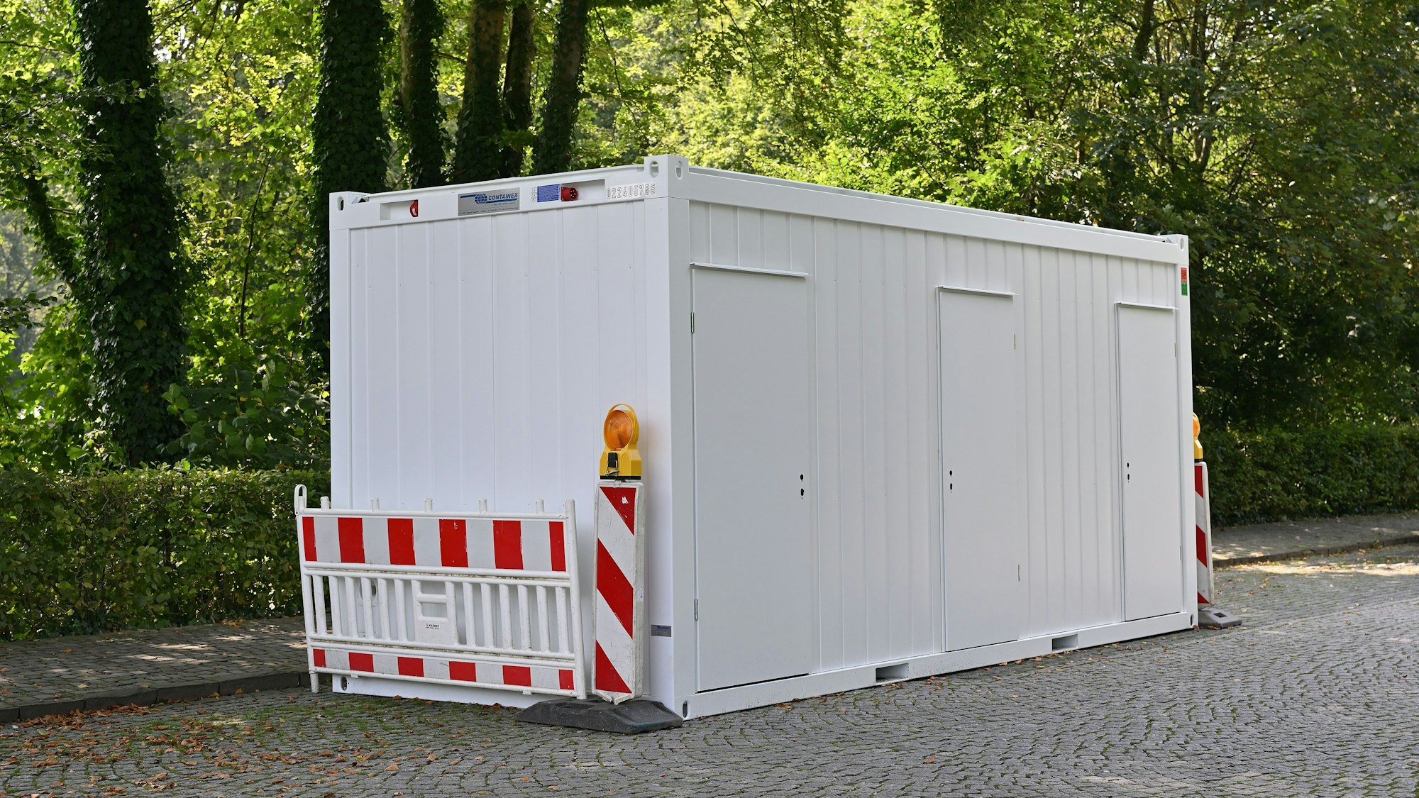 Ein weißer Toilettencontainer mit drei Türen steht mit Absperrbaken auf einem Platz.
