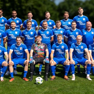 Die Mannschaft des TuS Dom-Esch posiert in blauen Trikots auf einem Rasenplatz. Im Hintergrund stehen unscharf Bäume.