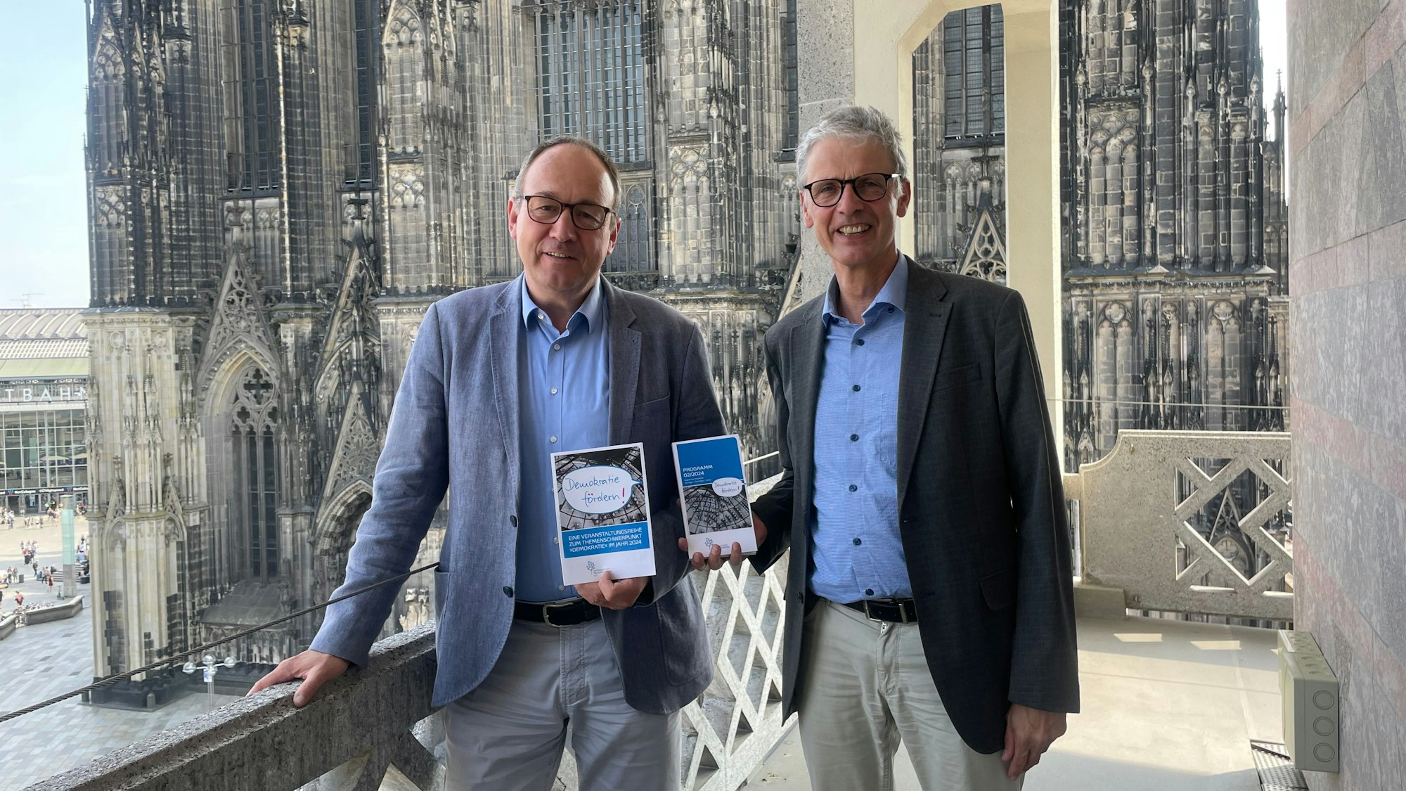 Zwei Männer stehen mit Programmheften in der Hand auf einem Balkon, hinter ihnen ist der Kölner Dom zu sehen.