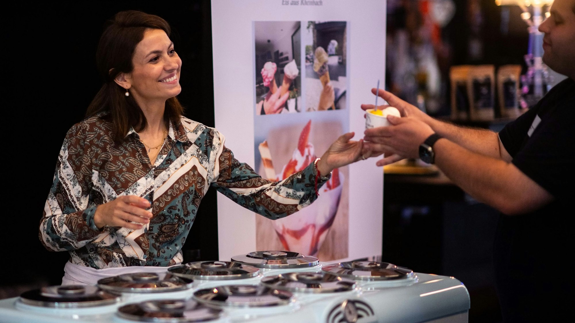 Das Bild zeigt eine Frau, die einen Becher Eis an einen Gast weitergibt.