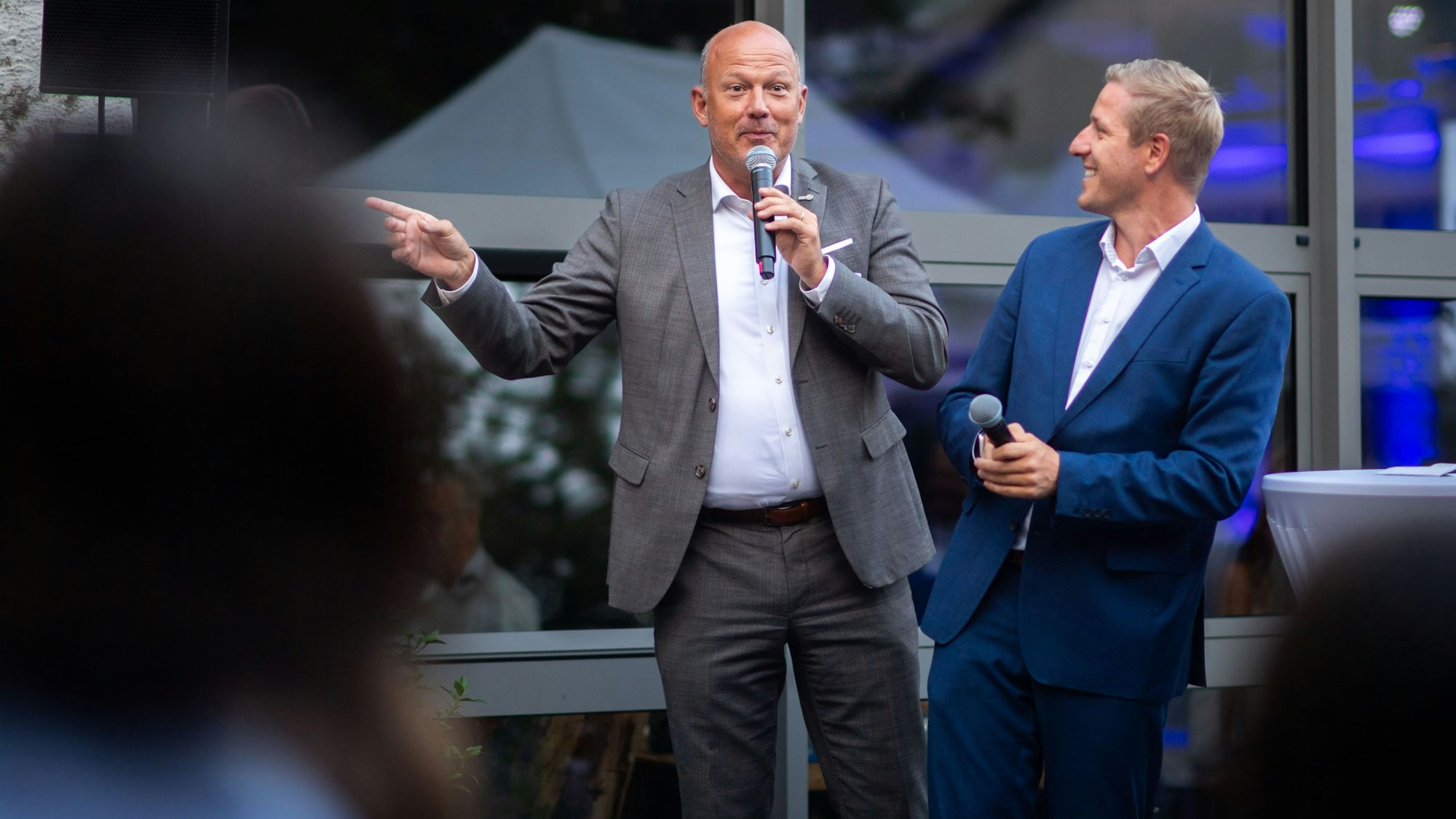 Das Bild zeigt die beiden Landräte auf der Bühne. Während Frank Rock gerade ins Mikrofon spricht, lächelt ihn sein Kollege Markus Ramers an.