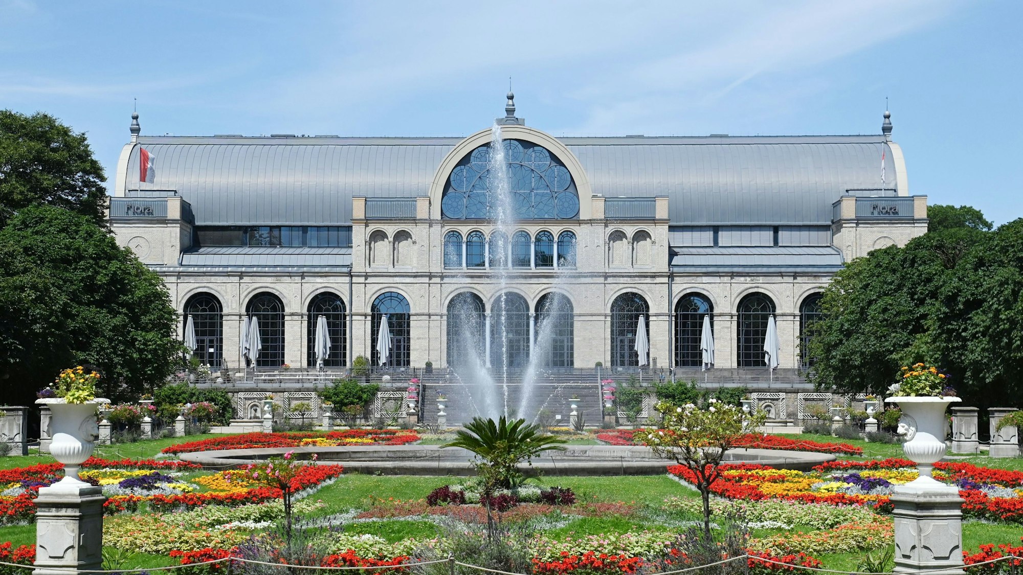 The main building of Flora Köln with a view of the flower beds and the fountain in front of it.