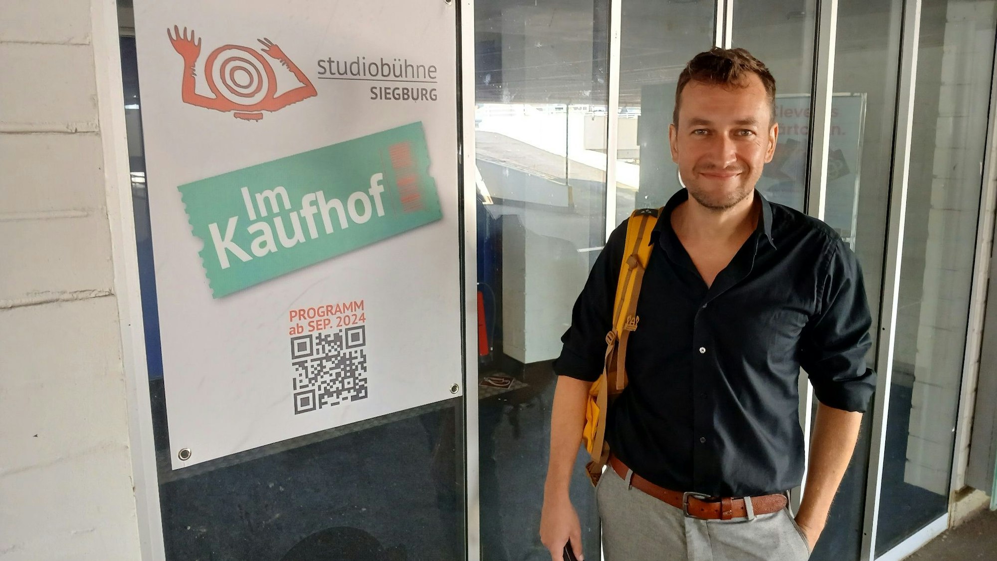 Ein Mann steht vor einer Glastür, daneben ein Plakat auf dem steht „Studiobühne Siegburg im Kaufhof“