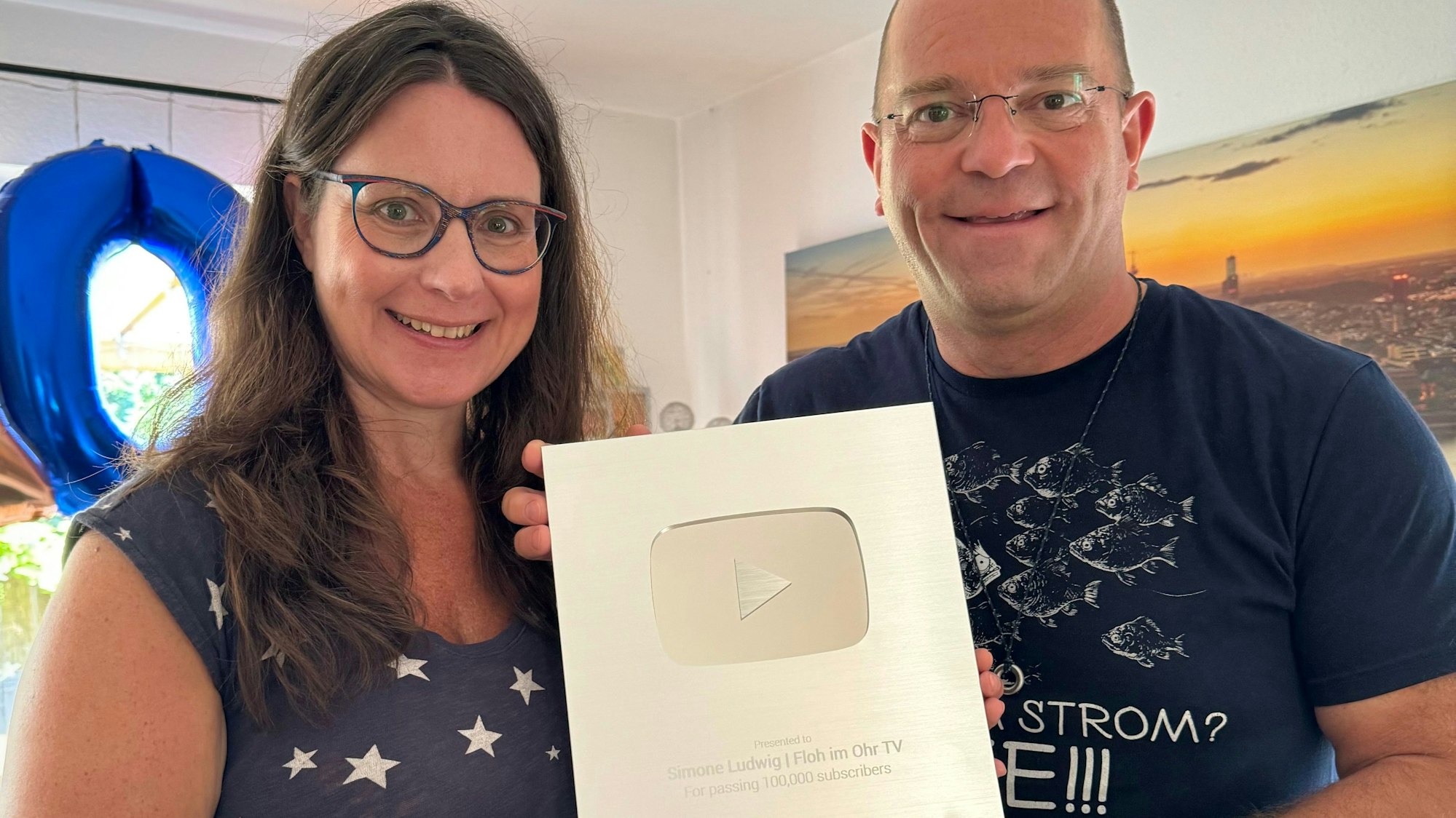 Eine Frau und ein Mann halten eine Auszeichnung in die Kamera und lächeln.