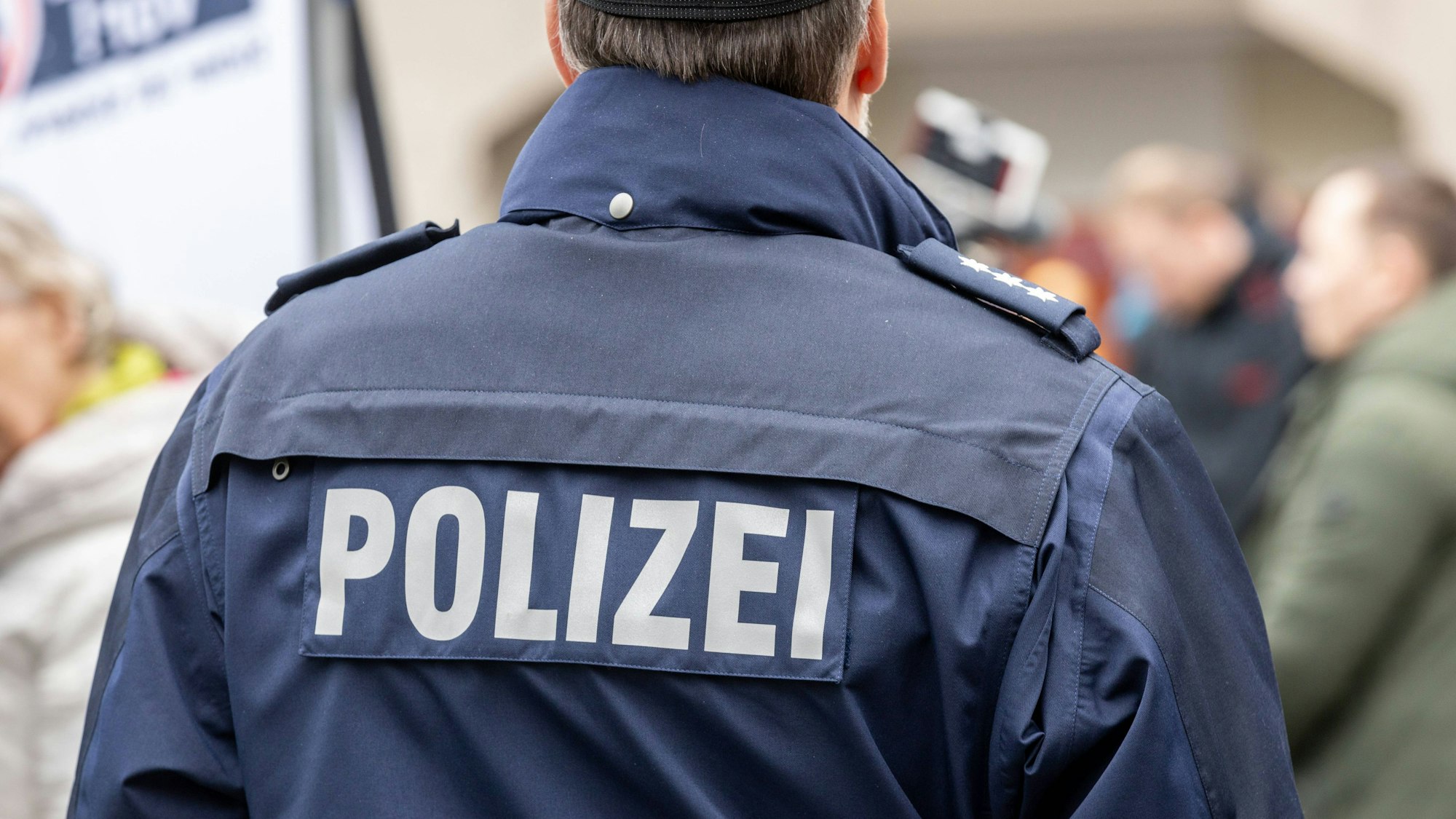 Ein Polizist ist von hinten zu sehen, auf seiner dunkelblauen Jacke steht Polizei.