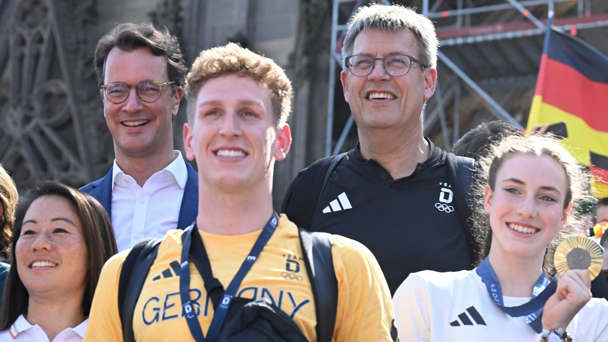 Vor dem Kölner Dom stehen Olympiamedaillen-Gewinner und Gewinnerinnen, unter ihnen Lukas Märtens (M) und Darja Varfolomeev (r), hinter ihnen DOSB-Präsident Thomas Weikert (oben r) und NRW-Ministerpräsident Hendrik Wüst. Darja Varfolomeev hält ihre Goldmedaille in der Hand, Lukas Märtens hat sie umhängen. Alle strahlen. Im Bildhintergrund weht eine Deutschlandflagge.