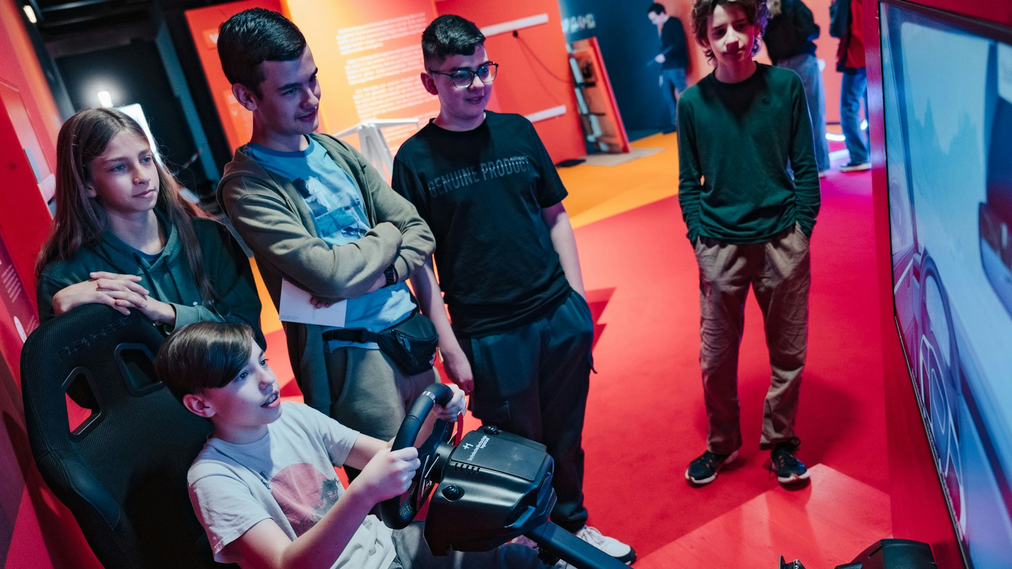 Eine Gruppe Jugendlicher vor einem großen Bildschirm, einer sitzt am Lenkrad eines Fahrsimulators.