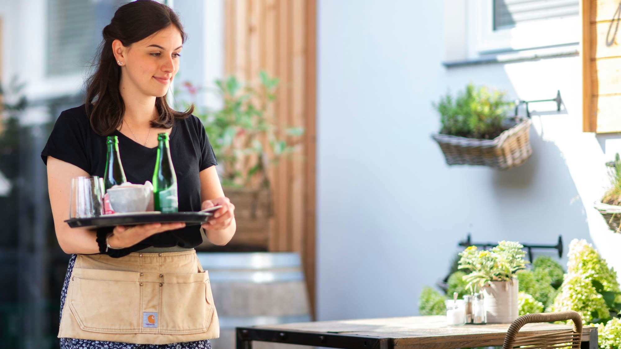 Das Bild zeigt eine Kellnerin, die im Außenbereich des Cafés auf einem Tablett Wasser und Kaffee transportiert.