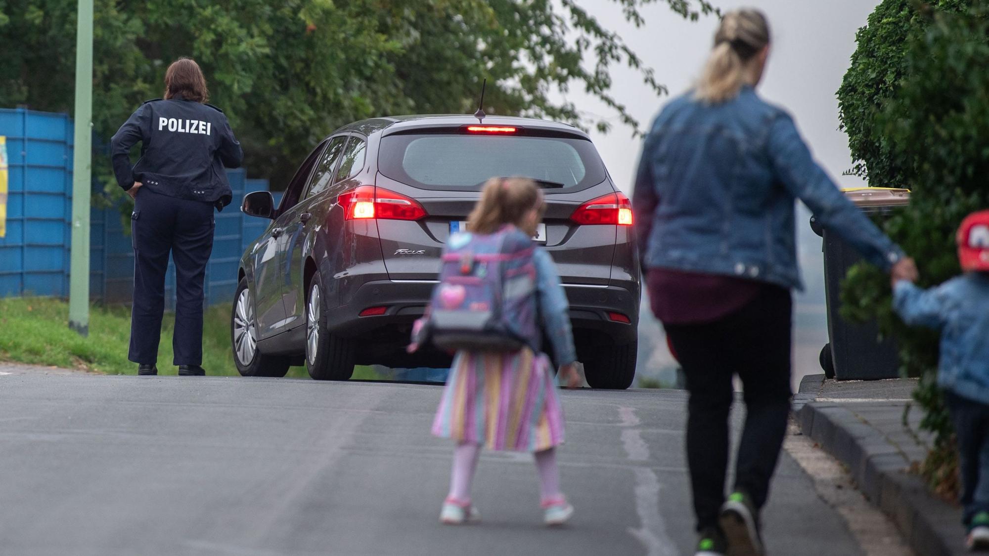 Eine Frau geht mit zwei Kindern über die Straße, im Hintergrund sind ein Auto und eine Polizistin zu sehen.