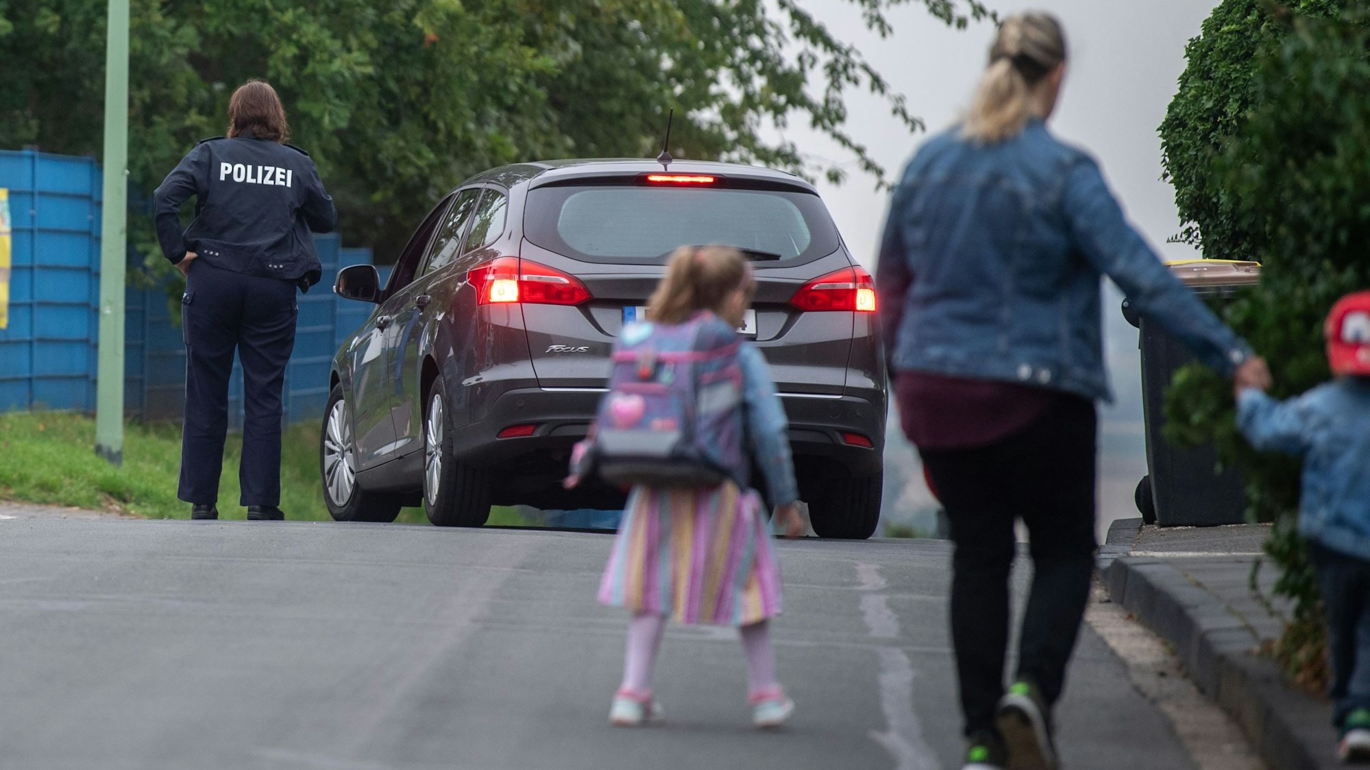 Eine Frau geht mit zwei Kindern über die Straße, im Hintergrund sind ein Auto und eine Polizistin zu sehen.